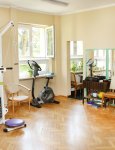 Wnętrze domu pomocy. Gabinet rehabilitacji ruchowej. Na drewnianym parkiecie stoją urządzenia rehabilitacyjne. Na kremowej ścianie trzy duże lustra. W głębi dwa okna z widokiem na ogród